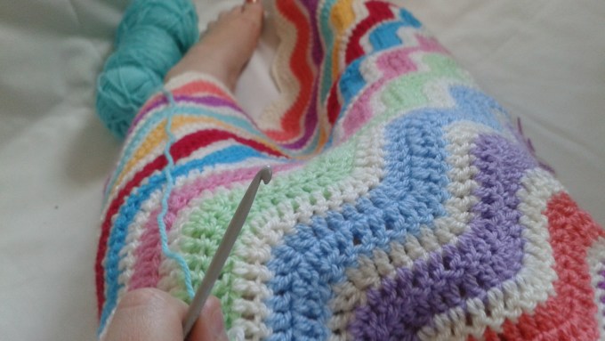 Crochet ripple blanket in progress, showing wavy stitches worked in soft yarn on crochet hook