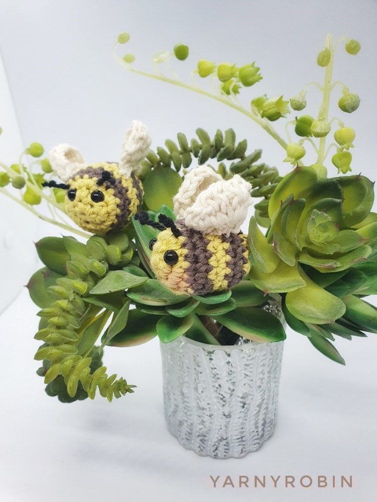 Two small crocheted amigurumi bees with cream wings nestled in a green succulent arrangement in a silver plant pot.
